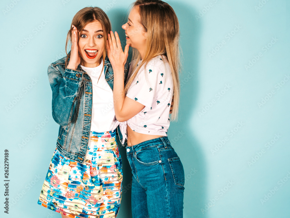 Two young beautiful blond smiling hipster girls in trendy summer casual clothes. Sexy women share secrets, gossip.Isolated on blue. Surprised face emotions Two young beautiful blond smiling hipster girls in trendy summer casual clothes. Sexy women share secrets, gossip.Isolated on blue. Surprised face emotions