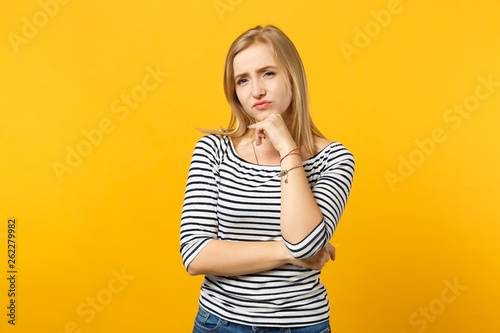 Puzzled young woman in striped clothes looking camera, put hand prop up on chin isolated on yellow orange wall background in studio. People sincere emotions, lifestyle concept. Mock up copy space.