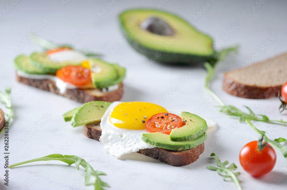 Toasts with avocado, cherry tomato and fried egg. The composition is supplemented with arugula greens. Light background. Toasts with avocado, cherry tomato and fried egg. The composition is supplemented with arugula greens. Light background.