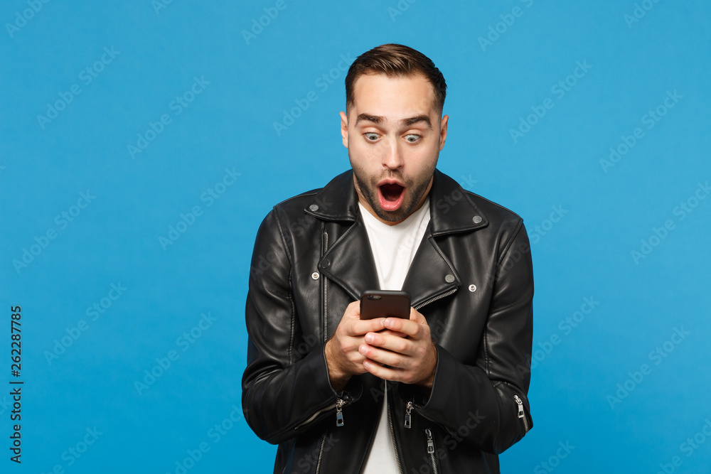 Handsome fun overjoyed excited young unshaven man in black leather jacket white t-shirt using mobile phone isolated on blue wall background studio portrait. People lifestyle concept Mock up copy space Handsome fun overjoyed excited young unshaven man in black leather jacket white t-shirt using mobile phone isolated on blue wall background studio portrait. People lifestyle concept Mock up copy space