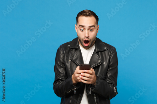 Handsome fun overjoyed excited young unshaven man in black leather jacket white t-shirt using mobile phone isolated on blue wall background studio portrait. People lifestyle concept Mock up copy space © ViDi Studio