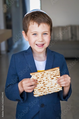 Cute Caucasian Jewish boy holding in his hands and taking a bite from a traditional Jewish matzo unleavened bread. Jewish Passover Pesach concept image. Vertical