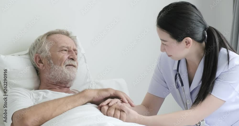 Nurse with senior patient in bedroom. Asian female nurse with bearded white male in bed. Holding hands and thumbs up. Senior lifestyle concept. Nurse with senior patient in bedroom. Asian female nurse with bearded white male in bed. Holding hands and thumbs up. Senior lifestyle concept.
