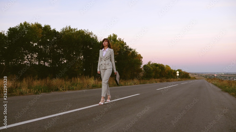 business woman with black briefcase walks in light suit and white high-heeled shoes goes outside city along asphalt with white markings, view from front