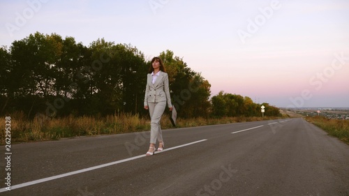 business woman with black briefcase walks in light suit and white high-heeled shoes goes outside city along asphalt with white markings  view from front