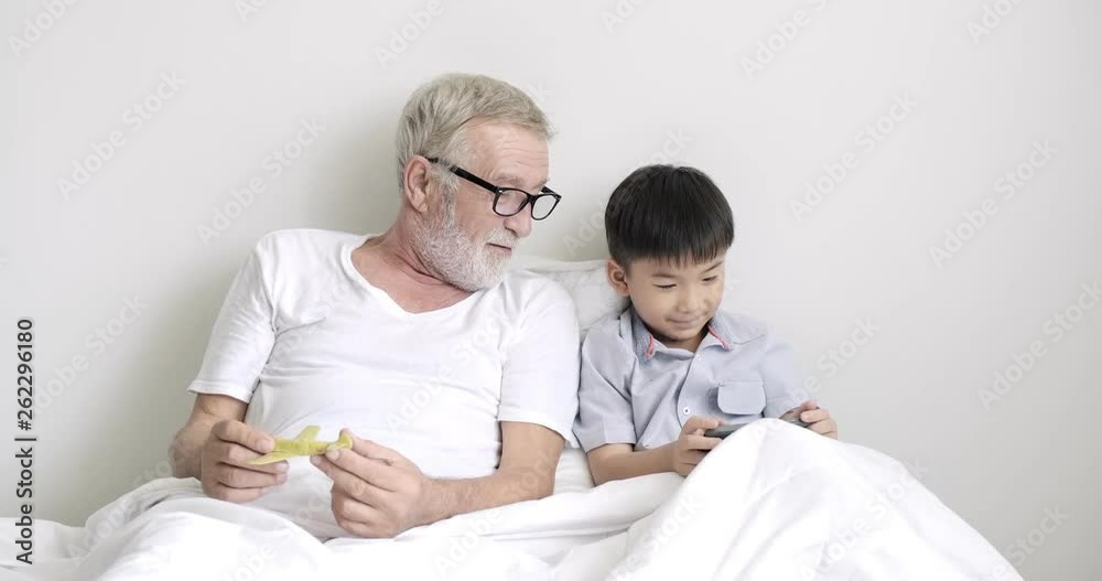 Senior grand father with grand son in bed together. White senior bearded man with young asian boy, playing mobile phone and toy, happy mood. Mix race, generation connection concepts. Senior grand father with grand son in bed together. White senior bearded man with young asian boy, playing mobile phone and toy, happy mood. Mix race, generation connection concepts.