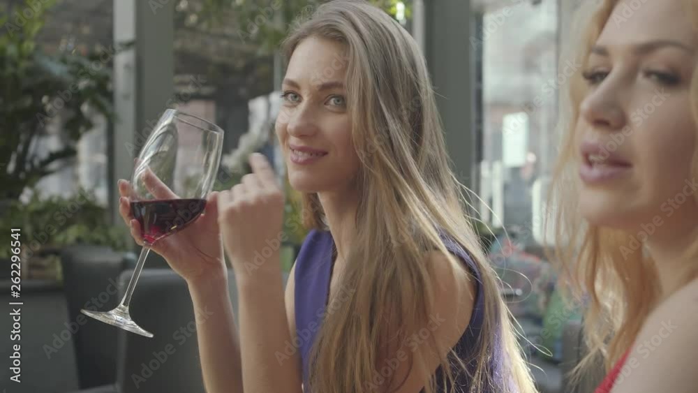 Two girlfriends sitting at the table in the cafe chatting and enjoying alcohol. Pretty elegant woman with long hair drinking red wine from the high glass. Curly blond lady telling story to the friend Two girlfriends sitting at the table in the cafe chatting and enjoying alcohol. Pretty elegant woman with long hair drinking red wine from the high glass. Curly blond lady telling story to the friend