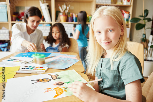 Adorable albino girl smiling and looking at camera while sitting at table and painting nice picture during lesson in art school