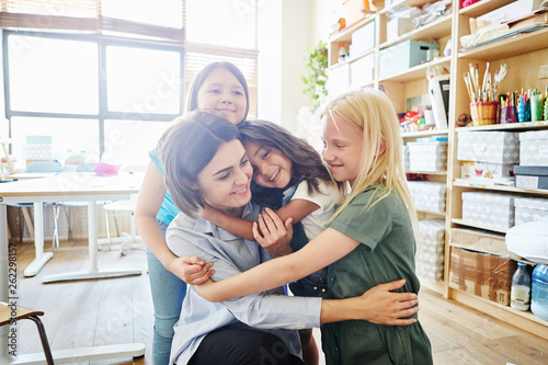 Cute girls smiling and embracing tenderly their young teacher in classroom at art school