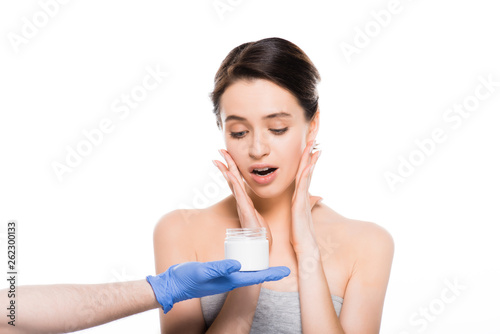cropped view of plastic surgeon in latex glove holding container with cosmetic cream near surprised girl isolated on white