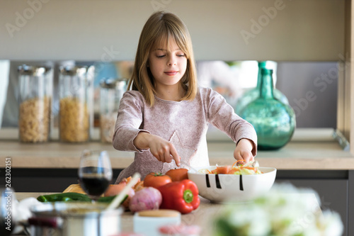Beautiful cute little girl preparing salad in a bowl in the kitchen at home.