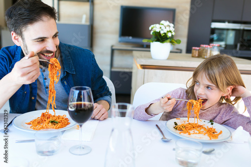 Handsome young father and her beautiful having fun while eating pasta with tomatoe sauce for lunch in the kitchen at home.