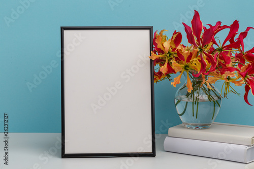 Composition from a decorative wooden frame and red and yellow flowers of Glorios in a glass vase on a white shelf on a blue background photo