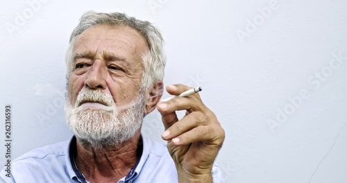 Senior man lighting up a cigarette with blue wall in background. Bearded white man. Enjoying his smoke.