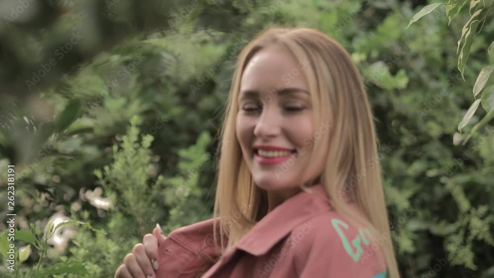 Beautiful blonde girl with red lips and blue eyes smiles sweetly to someone aside.She is in a botanical garden or a greenhouse