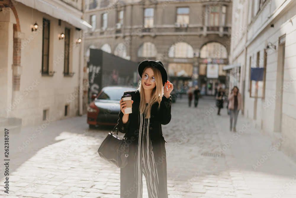 Fototapeta premium Young attractive lady with blonde straight hair, sunglasses, coat and hat is walking in the city with coffee to go