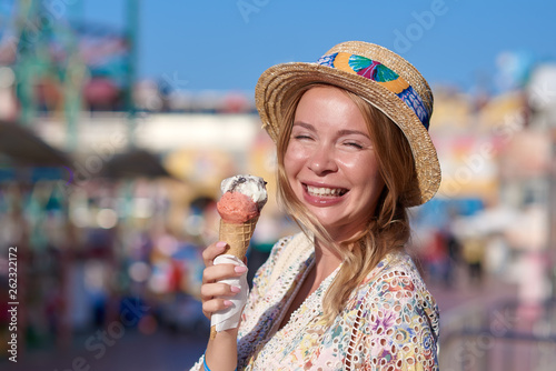 Young smiling woman in a wonderful dress and sunhat is eating an ice-cream in the Luna park. She is enjoying her holidays.