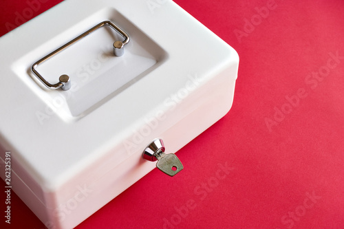 Close up of small white closed cashbox or money box with key in lock on dark pink background with copy space