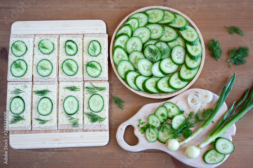  Light, spring sandwiches with cheese, fresh cucumber and dill, next to a tray with slices of cucumber and a board full of green spring vegetables