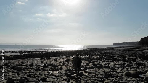 stunning 4k man walking on rocky beach towards the sea beautiful day drone over shoulder shot 