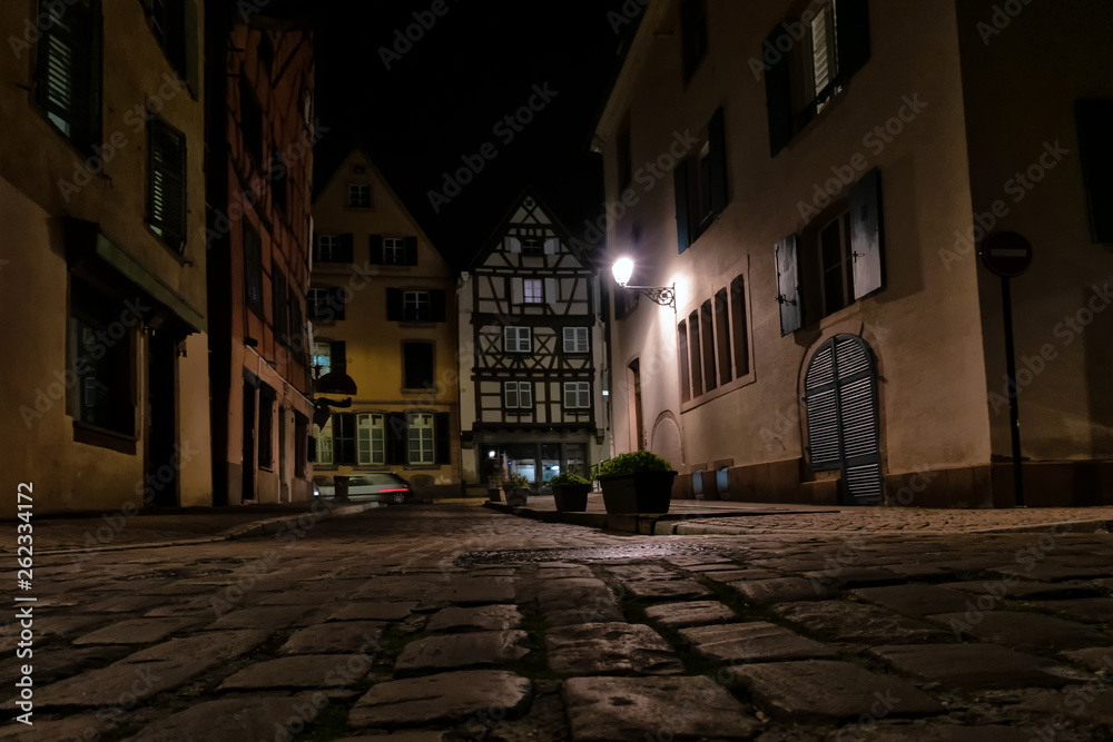 View of the street with old half-timbered houses. Evening time, shines a street lamp. View from below. In the foreground are pavement stones. In the background a car passes by. Background.