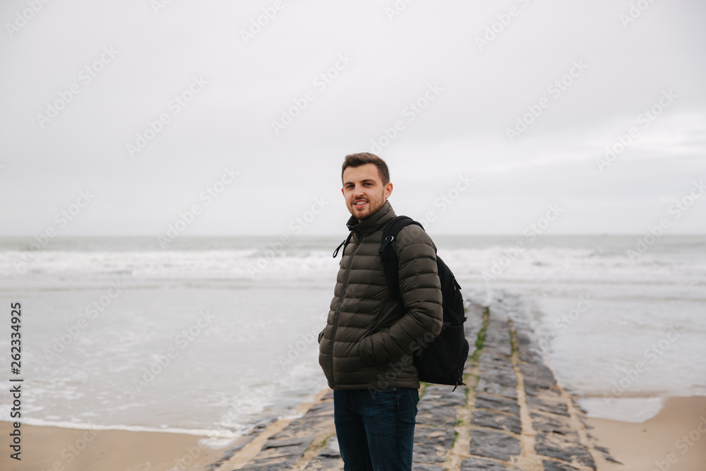 Male tourist walkin along the North Sea. Man with backpack stand on the coast in rainy weather. Beautiful sea Male tourist walkin along the North Sea. Man with backpack stand on the coast in rainy weather. Beautiful sea