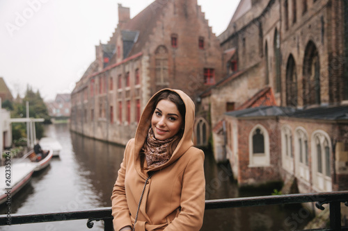 Portrait of beautiful woman in brown coat standing on the bridge in Bealgium, Brugge. Female put on a hood