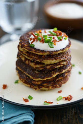Potato pancakes draniki served with sour cream, chives and bacon on a white plate. Dark wooden table, blue linen napkin, high resolution