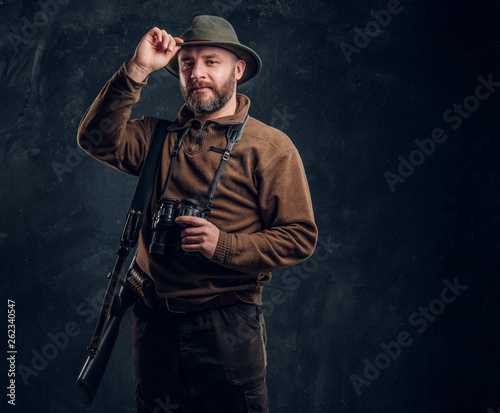 Bearded hunter with rifle and binoculars corrects his hat and looking at a camera. Studio photo against a dark wall background