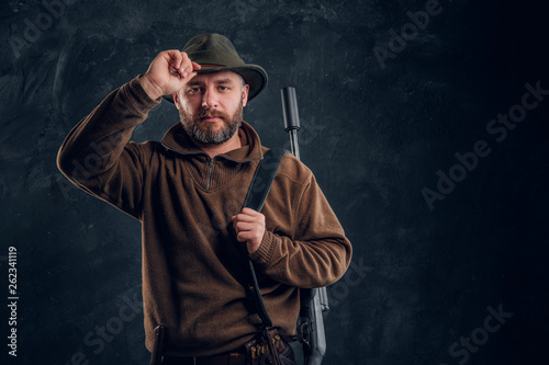 Portrait of a bearded hunter with rifle holding hand on hat and looking at a camera.
