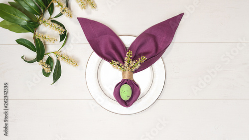 Beautiful Easter table setting with egg, purple napkin Easter Bunny, spring green tree branch on white wooden background. Flat lay, top view. Copy space.
