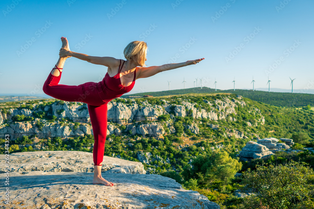 Yoga Fitness Woman Performing Lord of the Dance Pose (Natarajasana) with Stretched Foot and Hand. She is Standing at the Edge of Cliff Overlooking Mountain Range and Ocean in Far Distance.