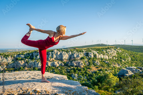 Yoga Fitness Woman Performing Lord of the Dance Pose (Natarajasana) with Bent Foot and Stretched Hand. She is Standing at the Edge of Cliff Overlooking Mountain Range and Ocean in Far Distance.