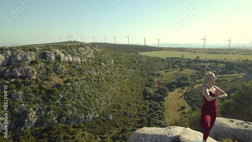 Aerial Footage of Mature Yoga Fitness Woman Performing Tree Pose (Vrikshasana ) on Cliff Edge of Ravine. Windmills in Background. Camera Moving Forward Towards Model.