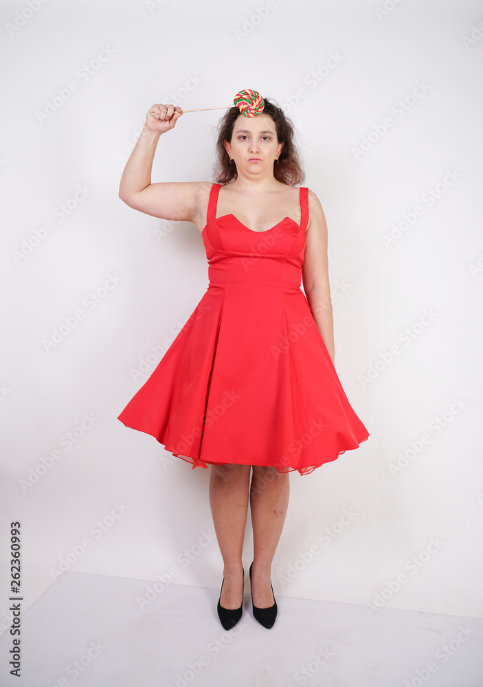 pretty chubby fashion girl wearing red pinup dress and posing with tasty colorful lollipop on white studio background