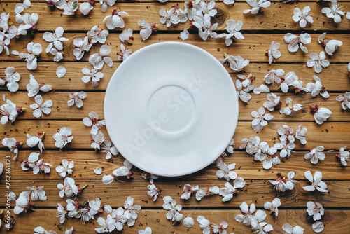 A white empty plate stands in the center of the table, surrounded by flowering apricot flowers on old brown background. Fine art, rustic, top view.