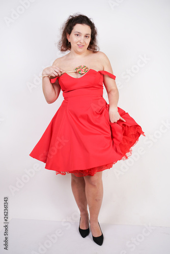 pretty chubby fashion girl wearing red pinup dress and posing with tasty colorful lollipop on white studio background
