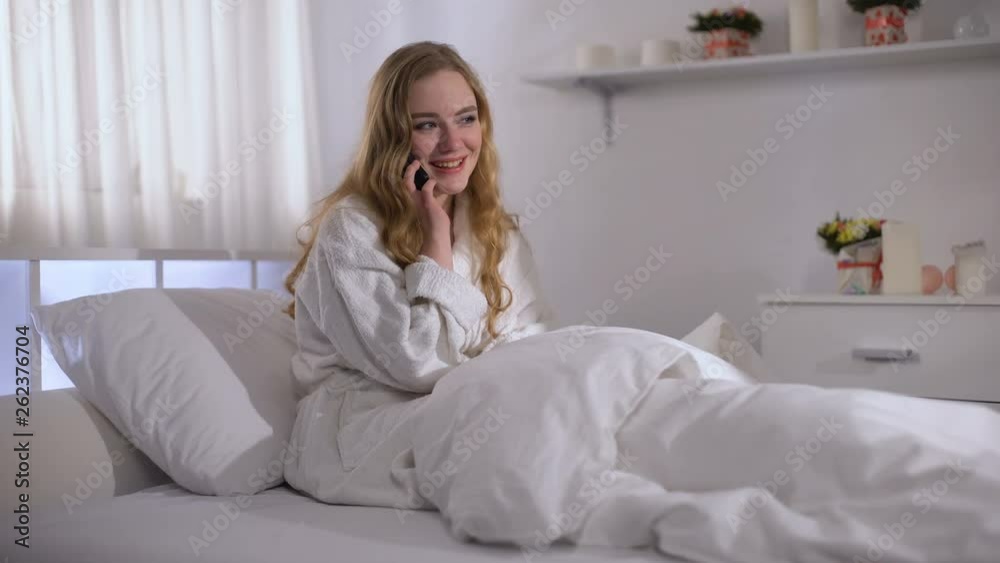 Cute girl happily talking on phone while lying in bed, planning day with friend Cute girl happily talking on phone while lying in bed, planning day with friend