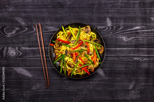Appetizing stir fried noodles with garlic sprouts, mushrooms, red pepper and chicken meat on a black rustic background of a wooden table. A popular Asian dish