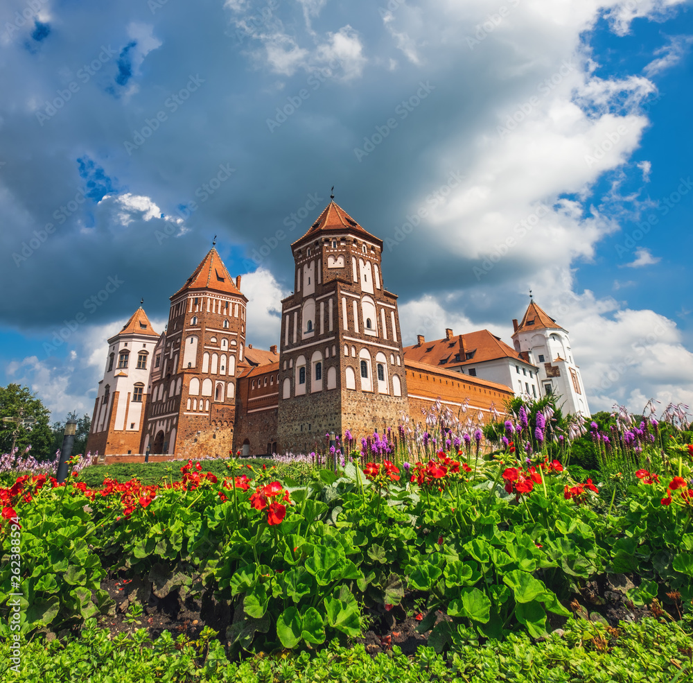 Multicolored flowers grow near the medieval castle in Mir, Belarus. Panoramic view of old castle under cloudy sky in summertime