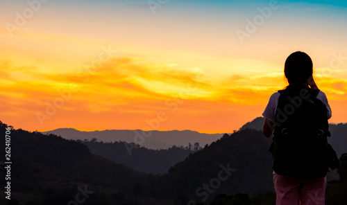 Back view of young woman watching beautiful sunset over mountain layer. Backpacker happy in travelling alone. Silhouette of tourist hiking to top of mountain. Adventure activity of active girl.