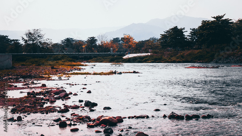 Bridge in Nakornnayok smalltown in Thailanda in the early morning with green trees along the river banks and big boulders reflecting in the water photo