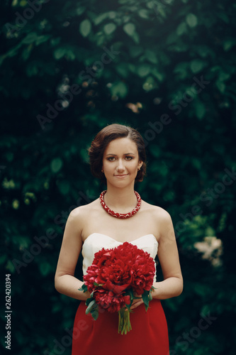 Gorgeous brunette bride in white and red dress holding flowers bouquet while posing in park, happy newlywed bride standing in a garden trees in the background, woman portrait