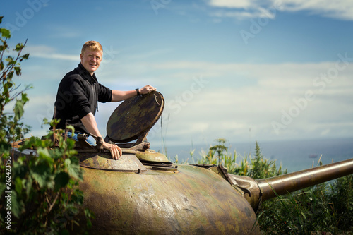 Redhead man in a black sweater stands in clear weather, leaning out of the old tank tower of the soviet T-55 tank on the Kamchatka Peninsula, Russia. Army and weapon, travel and adventure concept. photo