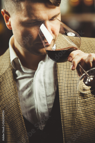 Sommelier smelling flavor of red wine in bokal on background of shelves with bottles in cellar. Male appreciating color, smell, quality and sediments of drink. photo