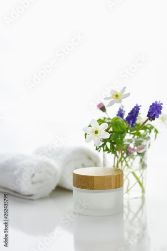 Weiße Handtücher neben einer Cremedose vor weißem Hintergrund und Blumen in einer Vase. 