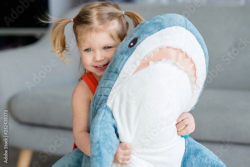 A little charming girl in a red dress is playing  with a big blue toy shark near gray sofa.