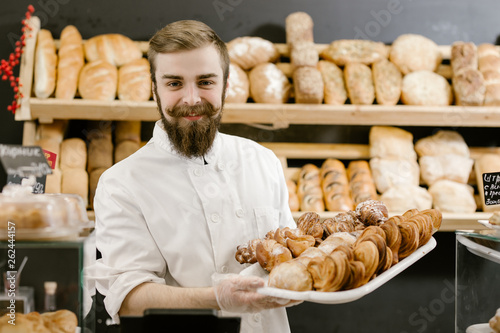 Charismatic baker with a beard and mustache stands with a tray with fresh pastries on the background of shelves with fresh bread in the bakery