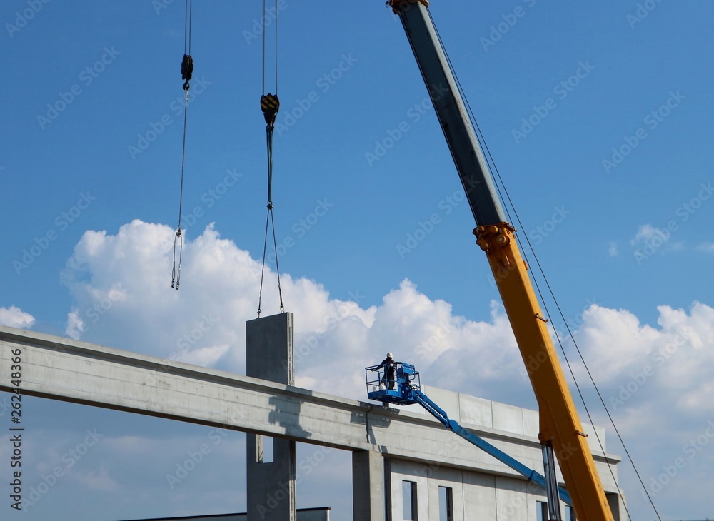 Worker on a cherry picker, together with  telescopic crane, installs a concrete block. It is  part  of an industrial building facade under construction