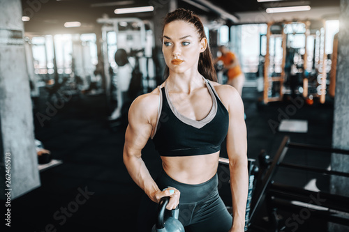 Pretty Caucasian female bodybuilder with ponytail lifting kettlebell. In background gym equipment. Get big or die trying.